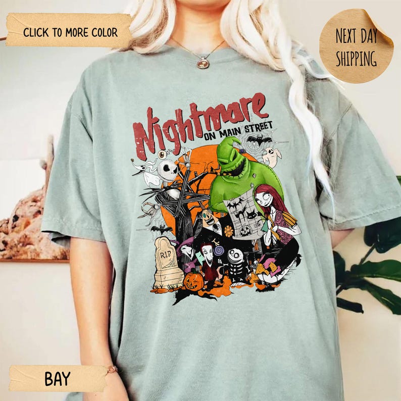 May include: A sage green t-shirt featuring a graphic print with the text Nightmare on Main Street. The design includes characters like Jack Skellington, Sally, and Oogie Boogie, along with a tombstone, a pumpkin, and bats against an orange moon.