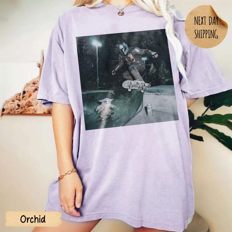 May include: Orchid-colored t-shirt featuring a graphic of The Mandalorian and Grogu skateboarding in a skate park. The image is printed on a soft, oversized tee. The shirt also has the text "Next Day Shipping".
