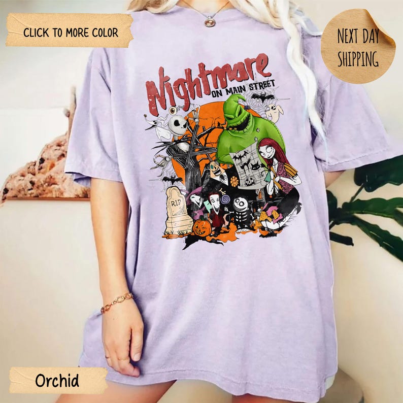 May include: A light purple t-shirt featuring a graphic print with the text Nightmare on Main Street. The design includes various characters from a movie, a large orange moon, a tombstone, a pumpkin, and bats against a dark background.