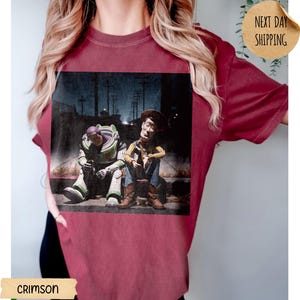 Pode incluir: Camiseta carmesim com um gr&aacute;fico desgastado de Buzz Lightyear e Woody de Toy Story. Os personagens s&atilde;o retratados sentados em uma cal&ccedil;ada em uma cena noturna. A camiseta tem gola redonda e mangas curtas.