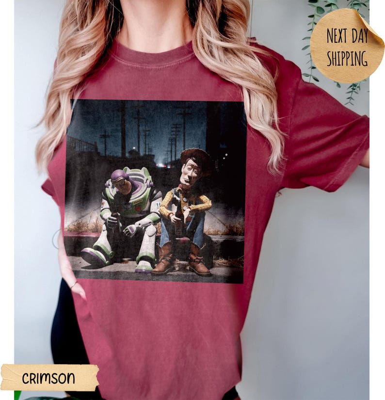 Pode incluir: Camiseta carmesim com um gr&aacute;fico desgastado de Buzz Lightyear e Woody de Toy Story. Os personagens s&atilde;o retratados sentados em uma cal&ccedil;ada em uma cena noturna. A camiseta tem gola redonda e mangas curtas.