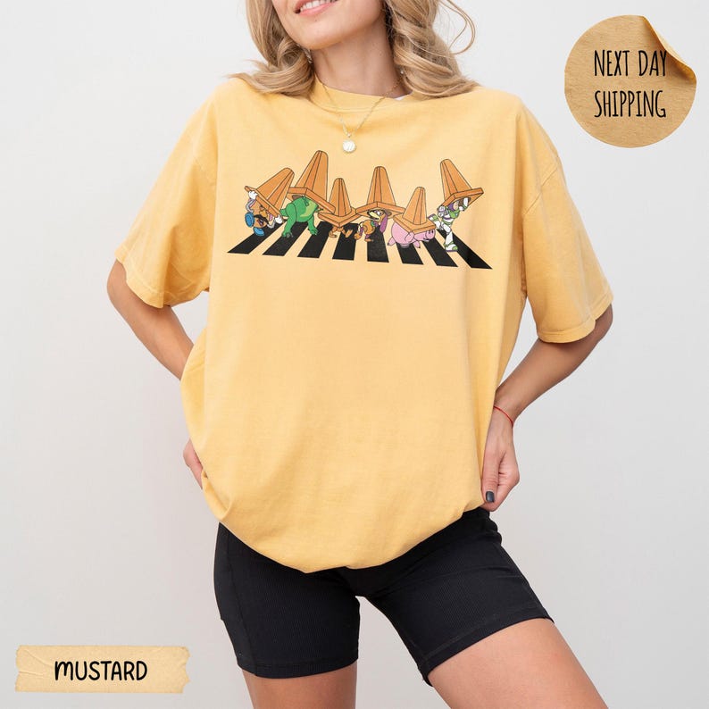 May include: A mustard yellow short-sleeve t-shirt featuring a graphic of cartoon characters walking across a black and white striped zebra crossing while wearing orange traffic cones on their heads. The shirt has a crew neckline and a loose fit.
