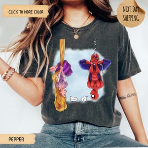 Op de afbeelding: Een antracietgrijs T-shirt met korte mouwen en een grafische print van een blond personage met lang haar dat aan een gouden vlecht hangt en een rood-blauwe superheld die aan een web hangt. De personages hangen ondersteboven met tekstballonnen.