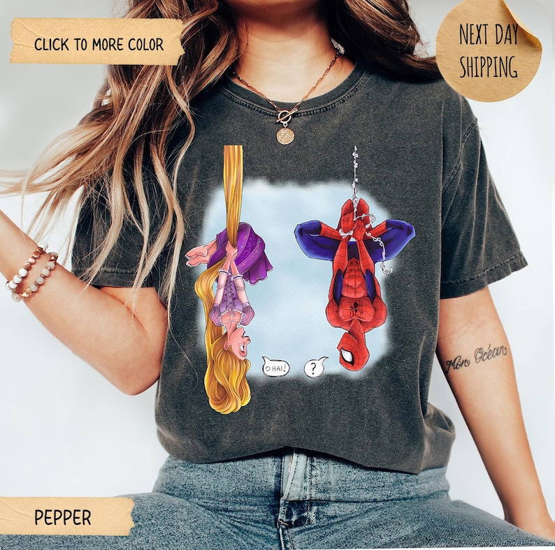 Op de afbeelding: Een antracietgrijs T-shirt met korte mouwen en een grafische print van een blond personage met lang haar dat aan een gouden vlecht hangt en een rood-blauwe superheld die aan een web hangt. De personages hangen ondersteboven met tekstballonnen.
