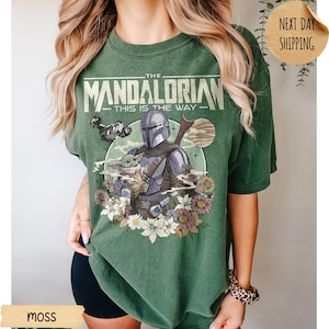 May include: Moss green t-shirt featuring a graphic of The Mandalorian and Grogu, with the text "THE MANDALORIAN - THIS IS THE WAY." The design includes a spaceship, flowers, and a moon. The shirt is a relaxed fit.