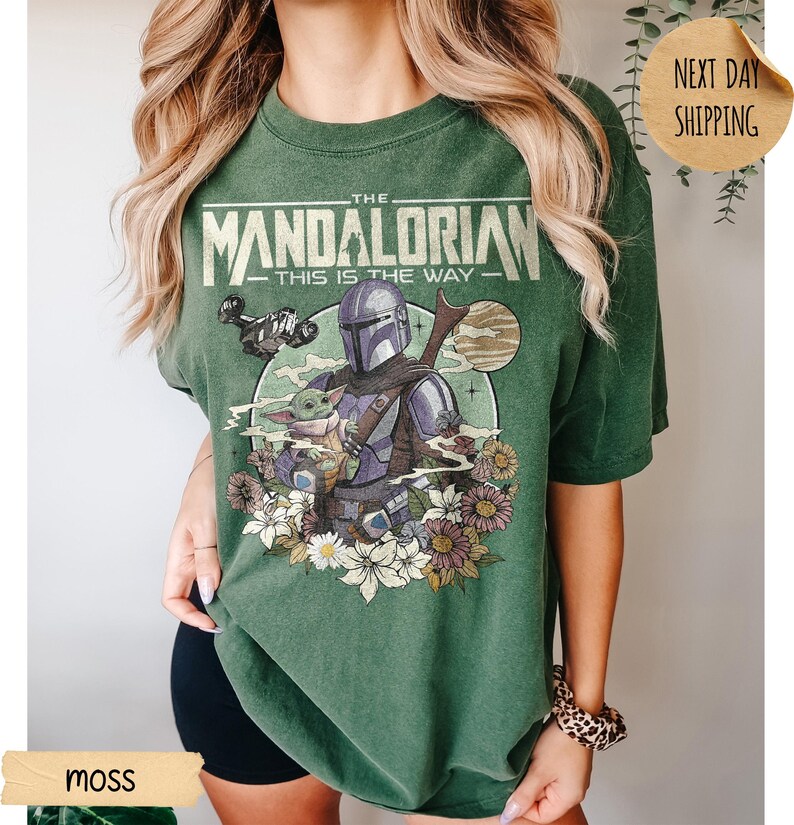 May include: Moss green t-shirt featuring a graphic of The Mandalorian and Grogu, with the text "THE MANDALORIAN - THIS IS THE WAY." The design includes a spaceship, flowers, and a moon. The shirt is a relaxed fit.