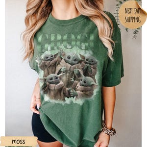 May include: A moss green short-sleeve t-shirt featuring a graphic print of five small green creatures with large ears and eyes. The text GROGU is printed in large letters above the creatures. The shirt has a crew neckline and a relaxed fit.