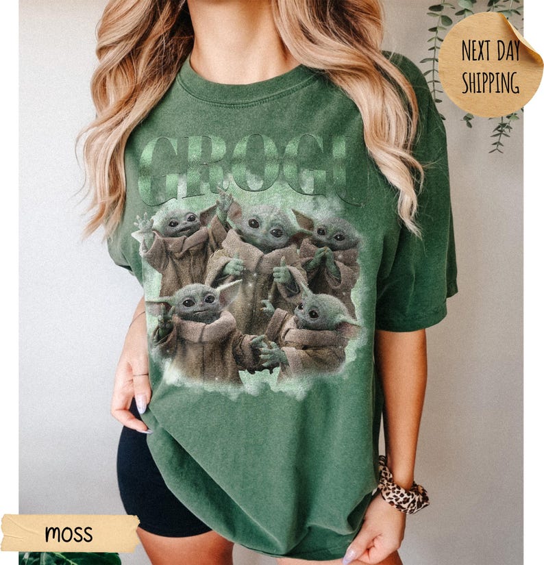 May include: A moss green short-sleeve t-shirt featuring a graphic print of five small green creatures with large ears and eyes. The text GROGU is printed in large letters above the creatures. The shirt has a crew neckline and a relaxed fit.