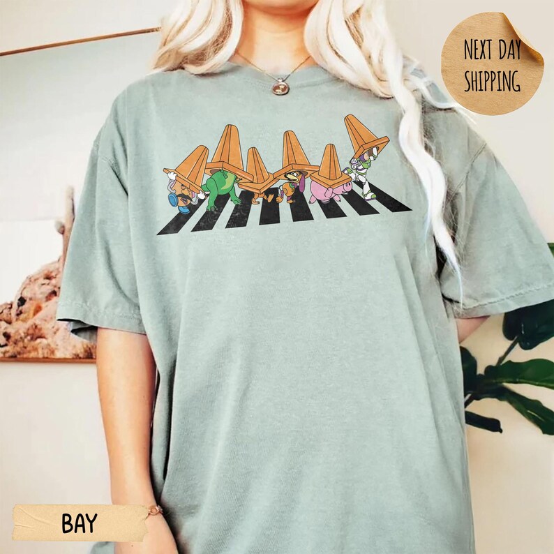 May include: A sage green short-sleeve t-shirt featuring a graphic of five cartoon characters walking across a black and white striped crosswalk, each wearing an orange traffic cone on their head. The design is centred on the chest of the shirt.