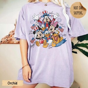 May include: A light purple short-sleeve t-shirt featuring a graphic print of Mickey Mouse, Minnie Mouse, Donald Duck, Daisy Duck, Goofy, and Pluto wearing patriotic hats. The design includes American flags, fireworks, and text America 250th Anniversary.