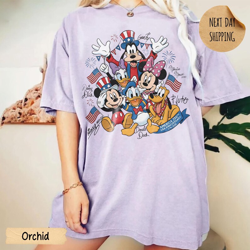 May include: A light purple short-sleeve t-shirt featuring a graphic print of Mickey Mouse, Minnie Mouse, Donald Duck, Daisy Duck, Goofy, and Pluto wearing patriotic hats. The design includes American flags, fireworks, and text America 250th Anniversary.