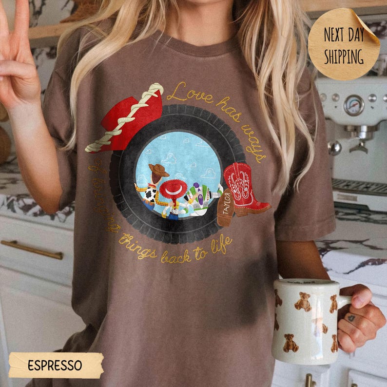 May include: A brown t-shirt featuring a graphic of a black tire framing a blue sky scene with cartoon characters. A red cowboy hat and a red cowboy boot are positioned on the tire. The text reads Love has ways and brings things back to life.