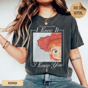 May include: A charcoal gray short-sleeve t-shirt featuring a graphic print of a cartoon character with red hair and a red hat inside a white square frame. The text reads EST 2026 I Knew It I Knew You with a small musical note symbol below.