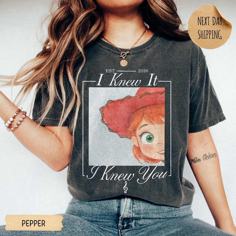 May include: A charcoal gray short-sleeve t-shirt featuring a graphic print of a cartoon character with red hair and a red hat inside a white square frame. The text reads EST 2026 I Knew It I Knew You with a small musical note symbol below.