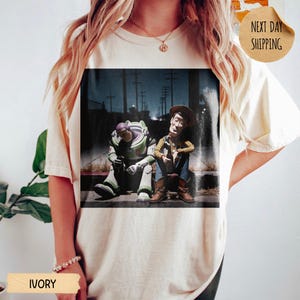 Pode incluir: Camiseta cor marfim com estampa gr&aacute;fica de Buzz Lightyear e Woody de Toy Story, sentados juntos. A imagem tem uma qualidade escura e cinematogr&aacute;fica. A camiseta tem um caimento folgado.