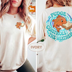 May include: Cream-coloured t-shirt with a cartoon fish and sandwich design. The front has a small graphic of the fish, while the back features a larger design with the text "Pudge Controls the Weather" in a circular pattern with blue and teal accents.