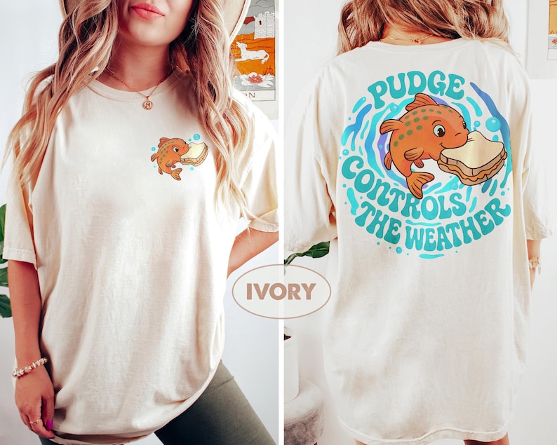 May include: Cream-coloured t-shirt with a cartoon fish and sandwich design. The front has a small graphic of the fish, while the back features a larger design with the text "Pudge Controls the Weather" in a circular pattern with blue and teal accents.
