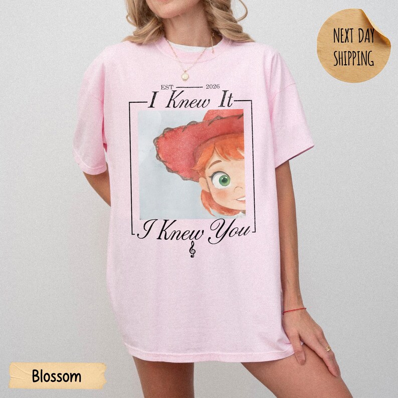 May include: A light pink t-shirt featuring a graphic of a cartoon character with red hair and a red hat inside a rectangular frame. The text reads "EST 2026", "I Knew It", and "I Knew You" with a small musical treble clef symbol below the text.