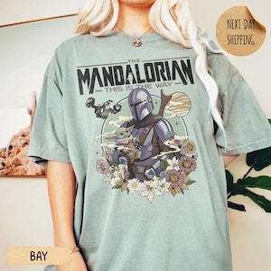 May include: A light green t-shirt featuring a graphic of The Mandalorian and Grogu surrounded by flowers. The text "THE MANDALORIAN - THIS IS THE WAY" is printed across the top. The shirt has short sleeves and a relaxed fit.