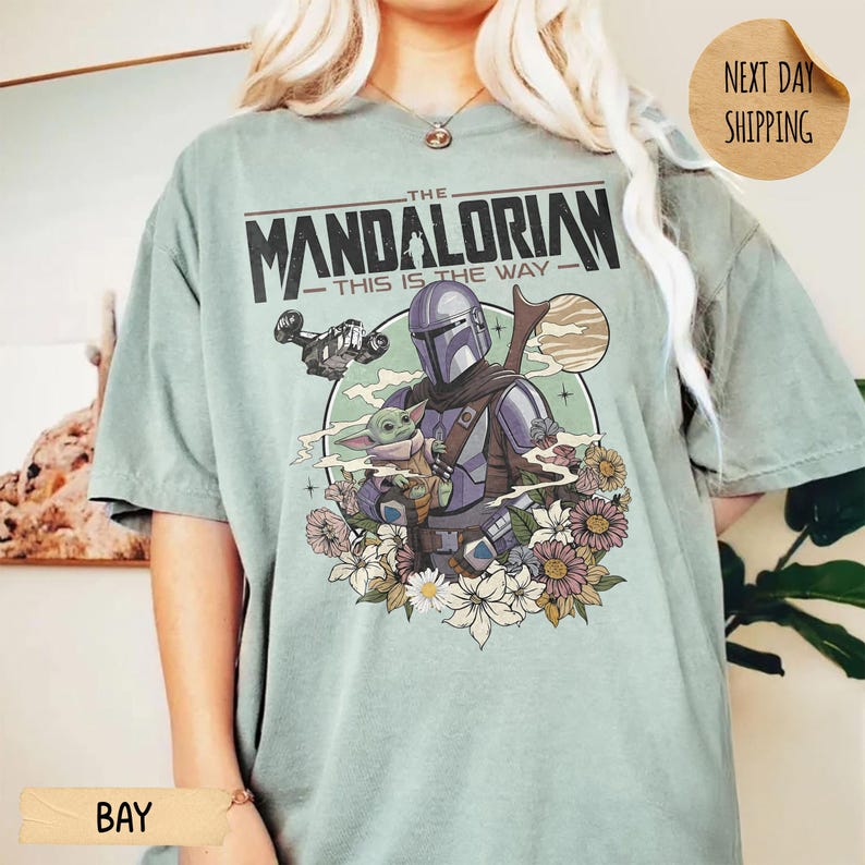May include: A light green t-shirt featuring a graphic of The Mandalorian and Grogu surrounded by flowers. The text "THE MANDALORIAN - THIS IS THE WAY" is printed across the top. The shirt has short sleeves and a relaxed fit.