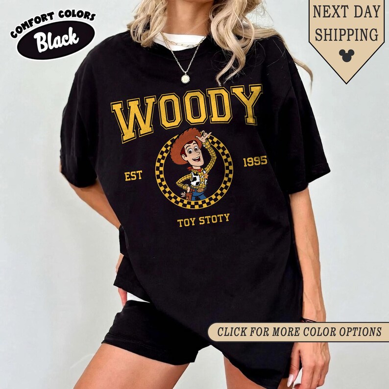 Puede incluir: Camiseta negra con un gr&aacute;fico de Woody. El dise&ntilde;o presenta la palabra "WOODY" en amarillo, una imagen de dibujos animados de Woody y el texto "EST 1995 TOY STOTY". La camiseta tambi&eacute;n tiene las palabras "COMFORT COLORS Black" y "CLICK FOR MORE COLOR OPTIONS".