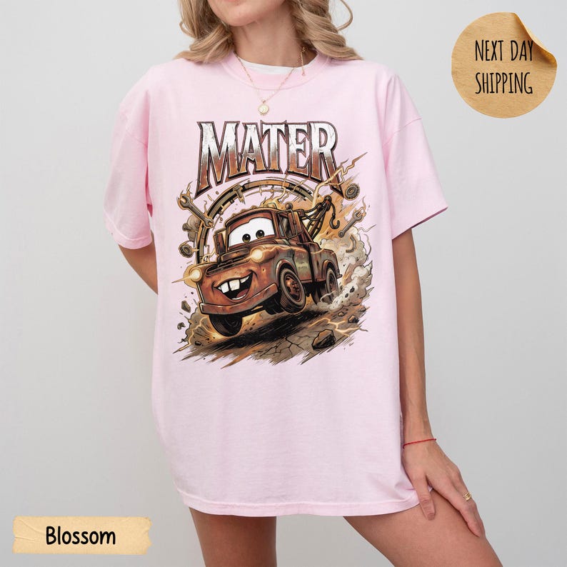May include: Pale pink t-shirt with a graphic of Mater from the film Cars. The design includes the word "MATER" above the character. The shirt has a relaxed fit.