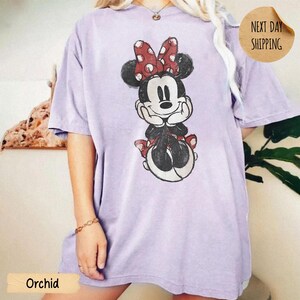 May include: A light purple short-sleeve t-shirt featuring a distressed graphic print of a cartoon mouse with a red and white polka dot bow. The mouse is depicted with hands resting under its chin, wearing a matching red and white polka dot skirt.