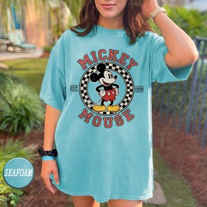 Puede incluir: Camiseta azul turquesa con un dise&ntilde;o de Mickey Mouse. El dise&ntilde;o incluye las palabras "Mickey Mouse" en rojo, un borde a cuadros y el a&ntilde;o "1928". La camiseta tiene mangas cortas y un corte holgado.