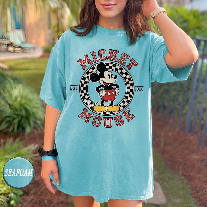 Puede incluir: Camiseta azul turquesa con un dise&ntilde;o de Mickey Mouse. El dise&ntilde;o incluye las palabras "Mickey Mouse" en rojo, un borde a cuadros y el a&ntilde;o "1928". La camiseta tiene mangas cortas y un corte holgado.