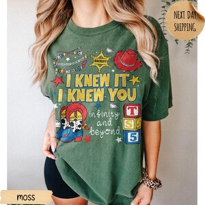 May include: A moss green t-shirt featuring a graphic print with the text I KNEW IT I KNEW YOU and infinity and beyond. The design includes a beaded bracelet, a yellow sheriff star, a red cowboy hat, cowboy boots, and stacked alphabet blocks.