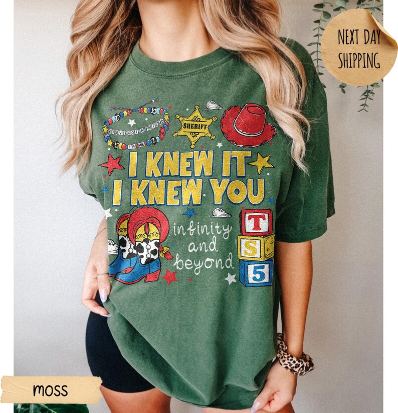 May include: A moss green t-shirt featuring a graphic print with the text I KNEW IT I KNEW YOU and infinity and beyond. The design includes a beaded bracelet, a yellow sheriff star, a red cowboy hat, cowboy boots, and stacked alphabet blocks.