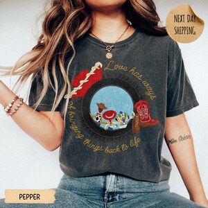 May include: A charcoal gray short-sleeve t-shirt featuring a graphic of a tire framing cartoon characters, a red cowboy boot, and a red hat. The text reads, Love has ways of bringing things back to life. The shirt has a vintage, washed-out appearance.