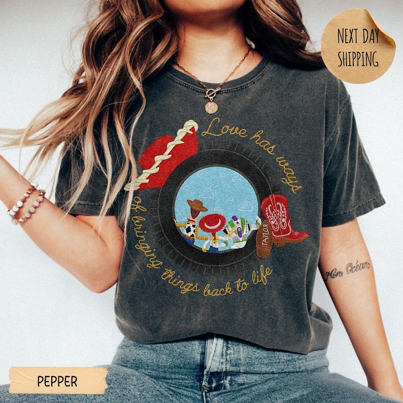 May include: A charcoal gray short-sleeve t-shirt featuring a graphic of a tire framing cartoon characters, a red cowboy boot, and a red hat. The text reads, Love has ways of bringing things back to life. The shirt has a vintage, washed-out appearance.