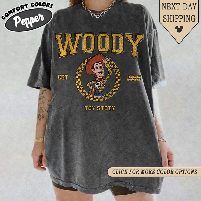 Puede incluir: Camiseta gris oscuro con un dise&ntilde;o de Woody de Toy Story. El dise&ntilde;o incluye la palabra "WOODY" en amarillo, el texto "EST 1995" y "TOY STOTY". La camiseta tiene los logotipos "COMFORT COLORS" y "Pepper".