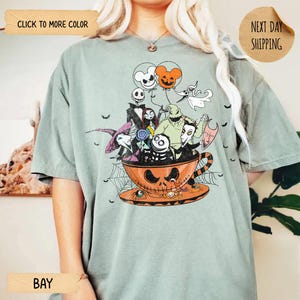 K&ouml;nnte beinhalten: Hellgr&uuml;nes T-Shirt mit einem Halloween-Motiv. Das Design zeigt Charaktere aus Nightmare Before Christmas in einer K&uuml;rbis-Teetasse, mit Luftballons und Flederm&auml;usen. Auf dem Shirt steht das Wort "BAY".