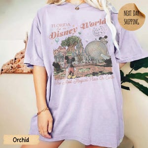 May include: Orchid-colored t-shirt featuring a graphic of Disney World in Florida. The design includes Mickey Mouse, the Tree of Life, and Spaceship Earth, with the text "Florida Disney World Est. 1971" and "The Most Magical Place On Earth."