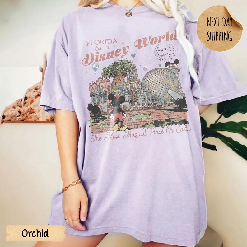 May include: Orchid-colored t-shirt featuring a graphic of Disney World in Florida. The design includes Mickey Mouse, the Tree of Life, and Spaceship Earth, with the text "Florida Disney World Est. 1971" and "The Most Magical Place On Earth."