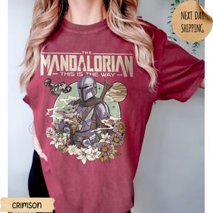 May include: Crimson t-shirt featuring a graphic of The Mandalorian and Grogu surrounded by flowers. The text on the shirt reads "THE MANDALORIAN - THIS IS THE WAY". The design includes a spaceship and a planet.