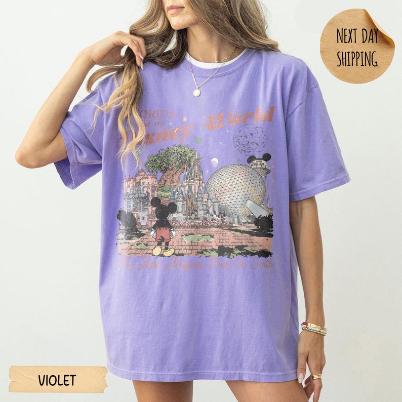 May include: Violet short-sleeved t-shirt featuring a graphic of Mickey Mouse in front of a theme park. The shirt has the words "World" and "Orlando" printed on it. The shirt is a relaxed fit.