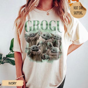 May include: An ivory short-sleeve t-shirt featuring a graphic print of five green creatures in tan robes. The text GROGU is printed in large green letters above the characters. The design includes a soft green watercolor-style background effect.