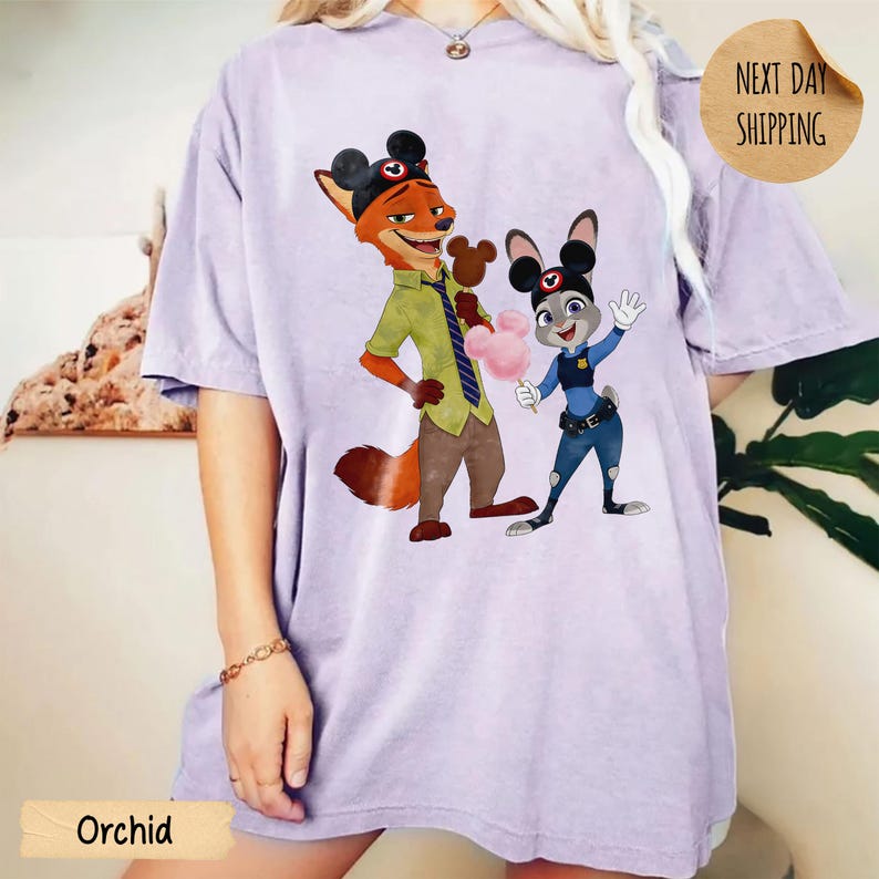 May include: Orchid-colored t-shirt featuring a graphic of Nick Wilde and Judy Hopps from Zootopia, each wearing Mickey Mouse ears. Nick holds a Mickey-shaped treat, and Judy holds cotton candy. The shirt has a relaxed fit.