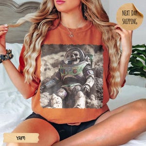 May include: A burnt orange crewneck sweatshirt featuring a graphic print of a skeleton wearing a space suit with a clear helmet. The design shows the figure sitting on a rocky, cratered surface. The shirt has a round neckline and short sleeves.