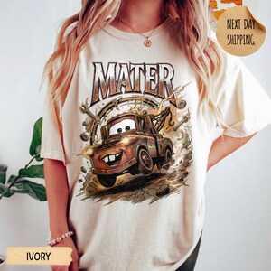 May include: Ivory t-shirt with a cartoon image of Mater from the film Cars. The design shows Mater smiling, with bolts and lightning around him, and the word "MATER" above. The shirt is a relaxed fit and made of soft material.
