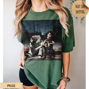 Pode incluir: T-shirt verde musgo com um gr&aacute;fico desgastado de Buzz Lightyear e Woody de Toy Story sentados em degraus. A imagem tem um visual vintage. A palavra "moss" &eacute; vis&iacute;vel. Corte folgado.
