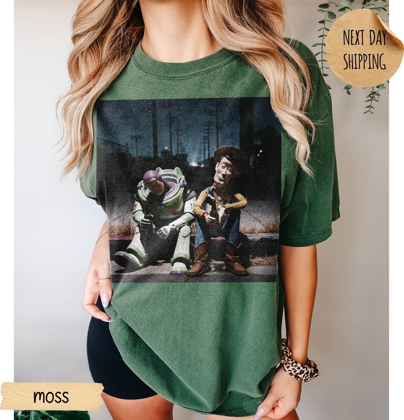 Pode incluir: T-shirt verde musgo com um gr&aacute;fico desgastado de Buzz Lightyear e Woody de Toy Story sentados em degraus. A imagem tem um visual vintage. A palavra "moss" &eacute; vis&iacute;vel. Corte folgado.