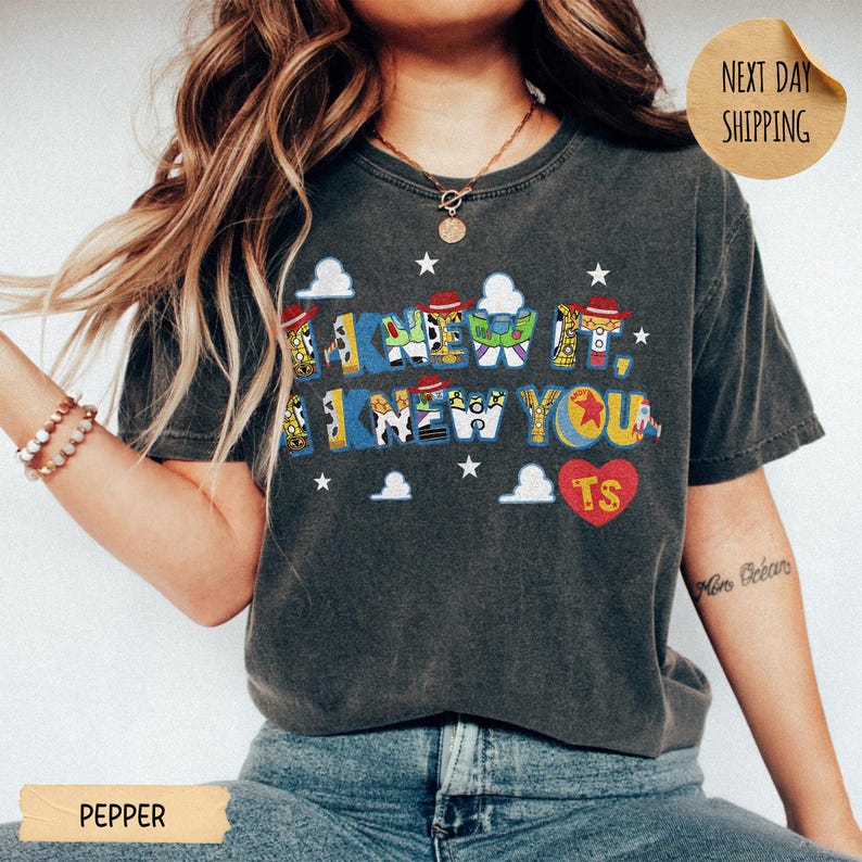 May include: A charcoal gray short-sleeve t-shirt featuring a graphic print with the text I KNEW IT, I KNEW YOU in blue letters filled with character patterns. The design includes small white stars, clouds, and a red heart with the letters TS inside.