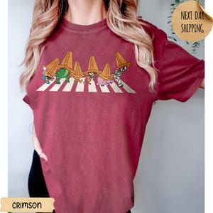 May include: A crimson short-sleeve t-shirt featuring a graphic print of six cartoon characters walking in a line across a white crosswalk, each wearing an orange traffic cone on their head. The design is centred on the chest of the garment.