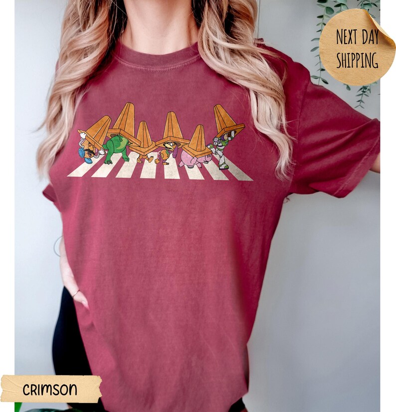 May include: A crimson short-sleeve t-shirt featuring a graphic print of six cartoon characters walking in a line across a white crosswalk, each wearing an orange traffic cone on their head. The design is centred on the chest of the garment.
