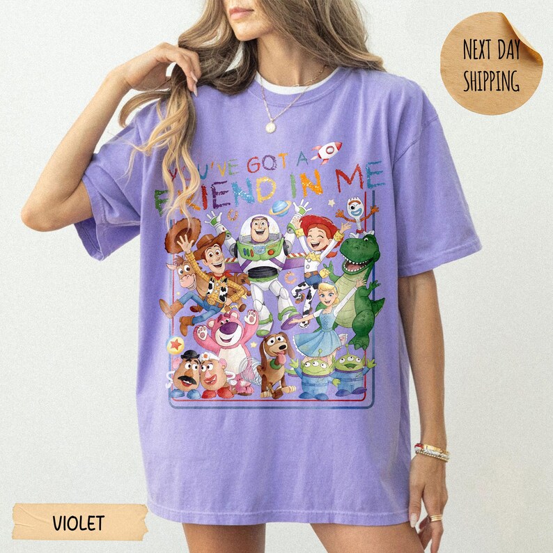 May include: A violet short-sleeve t-shirt featuring a colorful graphic print of various Toy Story characters including Woody, Buzz Lightyear, Jessie, and Rex. The text "YOU'VE GOT A FRIEND IN ME" is printed in multi-colored letters above the characters.