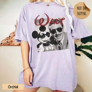 May include: A light purple t-shirt featuring a black and white image of Mickey Mouse and Walt Disney wearing sunglasses. The word "Walt" is printed in red above the image. The shirt has a relaxed fit.